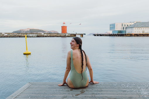 A woman enjoying thr winter swimming time relaxing on the wooden terrace with view of the ocean and the beautiful lighthouse in Alesund