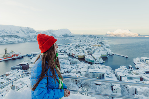 Side view of female in red hat and blue jacket admiring a view of Alesund during winter time with view of the scenic mountain and the sea during winter sunset
