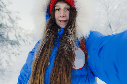 Smiling female with long hair in blue jacket making selfie in the majestic snowcapped forest with mountains view catching snow during sunset in Scandinavia