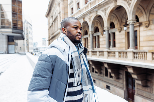 Young African man in casual white and blue clothes, striped sweater. Winter season street portrait with snow. Lifestyle, city life concept. Side view, copy space