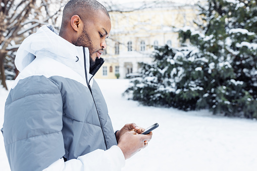 Young African American man in casual white clothes typing message, texting on phone, in park or street outdoor. Winter season with snow. Side view, copy space