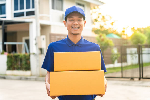 istock Handsome asian smiley delivery man wearing blue uniform holding parcel cardboard boxes in front of customer house and he so happy with his job. 1455284231