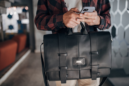 One man, unrecognizable male holding a modern leather bag and using smart phone.