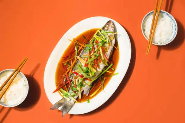 Photo of Cantonese-Style Recipes for  Chinese New Year festive table.  Sea bass or Spigola fish steamed with ginger, red peppers in soy sauce served white rice. Steamed fish - a symbol of prosperity and wellness in China