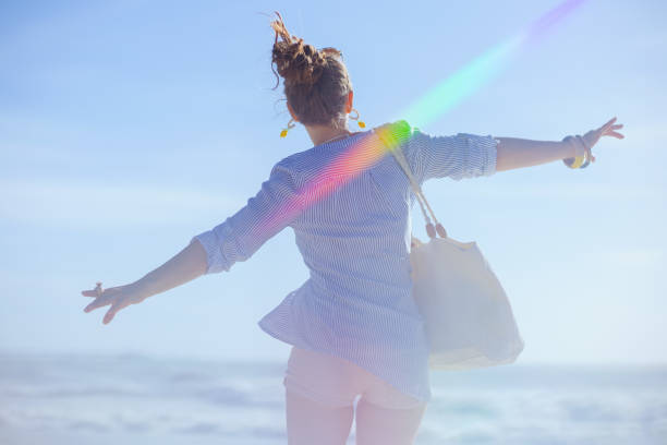 Seen from behind middle aged woman with white straw bag rejoicing at the beach.