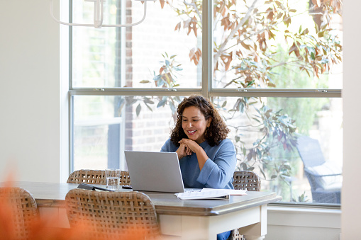 The mid adult woman sits in the dining room and uses her laptop for a video call with her family.