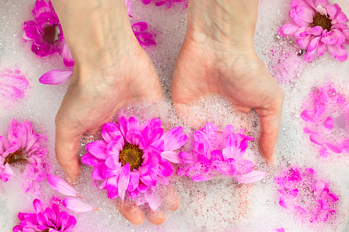 Close-up of female hands in bubble bath full of flowers and petals. Concept of body care, spa skin, natural cosmetics, purity and wellbeing. Top view, flat lay.