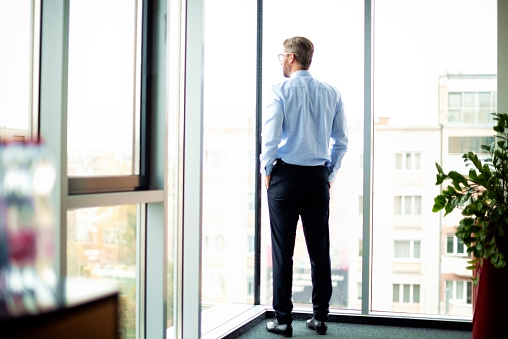Rear view of thinking businessman looking out of office over city. Confident man wearing glasses and business casual.