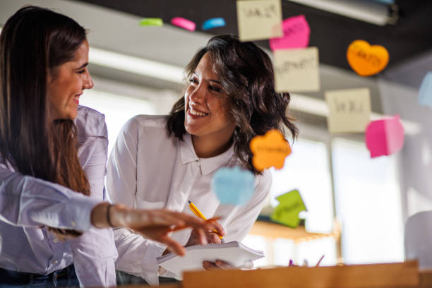 istock Two creative women brainstorming ideas and putting colorful sticky notes on a glass wall 1455296707
