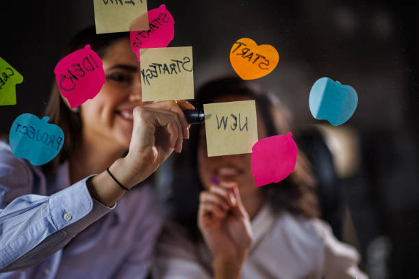 istock Two creative women brainstorming ideas and putting colorful sticky notes on a glass wall 1455296710