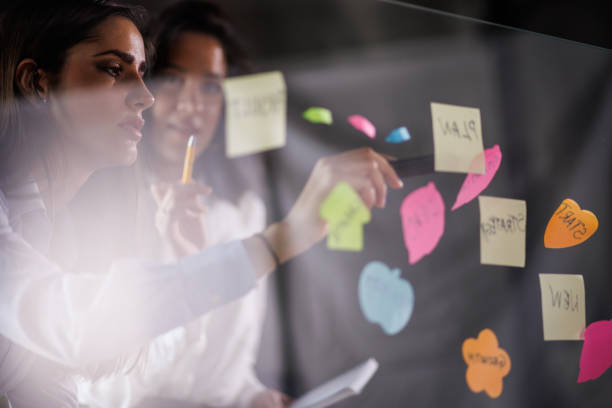 istock Two creative women brainstorming ideas and putting colorful sticky notes on a glass wall 1455296719