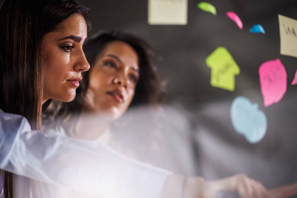 istock Two creative women brainstorming ideas and putting colorful sticky notes on a glass wall 1455296722