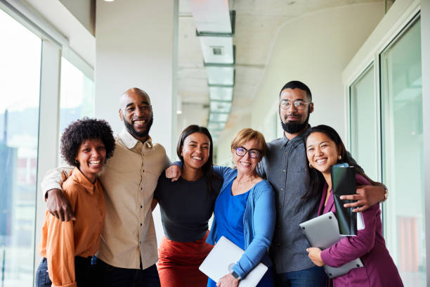 Smiling businesspeople standing arm in arm in an office hall Portrait of a group of diverse businesspeople smiling while standing arm in arm together in an office corridor multi ethnic group stock pictures, royalty-free photos & images