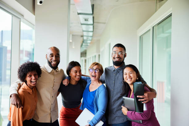 Laughing businesspeople standing arm in arm in an office hallway Portrait of a group of diverse businesspeople laughing while standing arm in arm together in the corridor of a modern office professional people laughing stock pictures, royalty-free photos & images