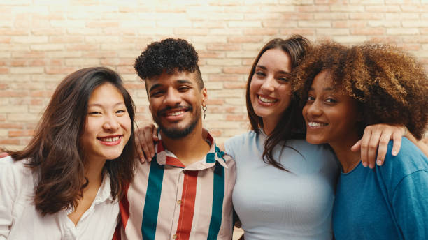 istock Happy, lovely multiethnic young people posing for the camera on summer day outdoors. Group of friends hugging tightly smiling at camera while standing on brick wall background 1455298467