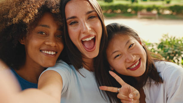 istock Happy, lovely multiethnic young women posing on cellphone camera on summer day outdoors. group of friends hugging each other smiling at the camera while standing on path in the park 1455298760