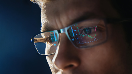 Close up view of focused trader wears eyeglasses looking at pc screen with stock market charts reflection, working online late night. Man analysing crypto market. Selective focus on eye. Copy space.