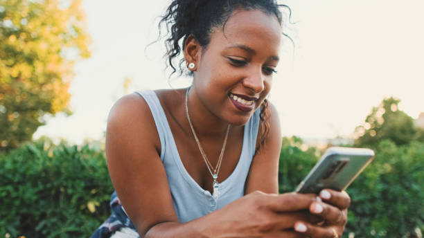 istock Laughing young mixed race woman sitting on park bench using cellphone 1455299807