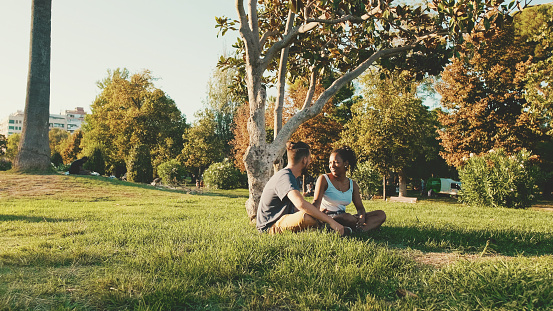 Happy smiling interracial couple talking while sitting on the grass in the park. Backlight