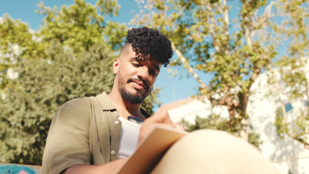 istock Young male student wearing an olive colored shirt takes notes in notepad while sitting on bench outside the university 1455300463