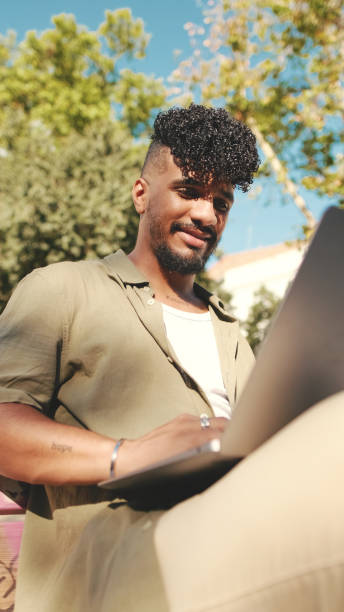 istock Close up, young male student wearing an olive colored shirt is working on pc laptop while sitting on bench outside the university 1455300545