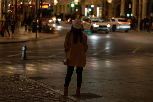 Rear view of young woman looking at night city lights, wearing brown jacket and hat