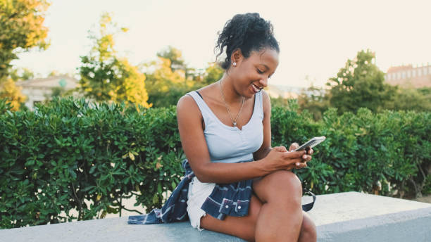 istock Laughing young mixed race woman sitting on park bench using cellphone 1455300782