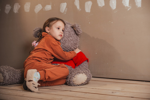 a little sad girl sits on the floor in an empty room, she hugged a huge teddy bear and looks thoughtfully somewhere