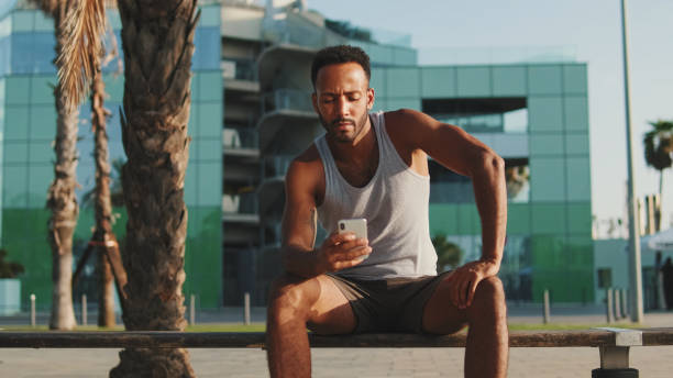 istock Young bearded male athlete is resting after training, sitting on bench, using smartphone 1455304634