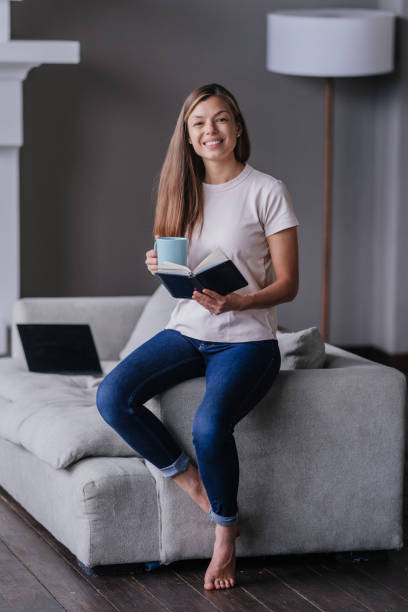 вертикальная съемка смайлика брюнетки молодой женщины в белой фут�болке держит чашку кофе и дневник смотрит в камеру зубастыми улыбками в г - book reading white women стоковые фото и изображения