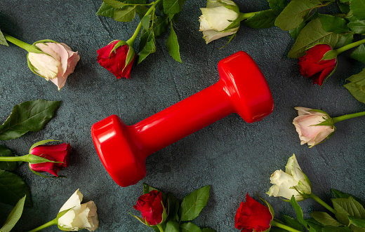 Red dumbbell as a love gift for Valentine's Day, marriage proposal engagement, birthday, anniversary, wedding. Healthy fitness, gym workout flat lay composition with roses and decorative hearts.