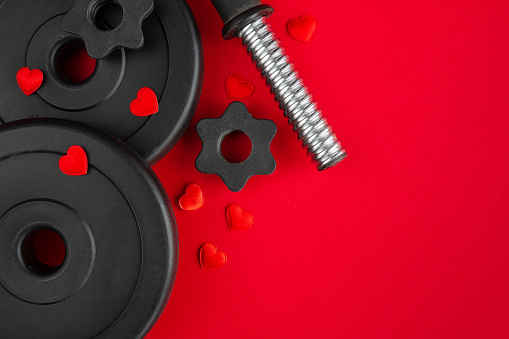 Dumbbells barbell weight plates and decorative hearts. Love gift for Valentine's Day, marriage proposal engagement, anniversary or wedding. Healthy fitness lifestyle composition. Gym workout and sport training flat lay concept with copy space on red background.