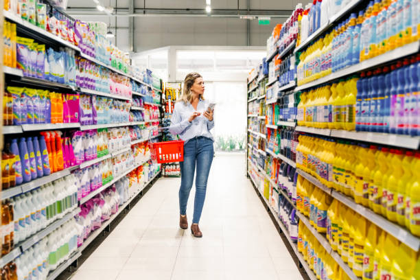 Woman customer with mobile phone buying groceries in store Full length of a mid adult woman with shopping basket and phone walking in supermarket aisle. Female customer with mobile phone buying groceries in store. holding shopping basket stock pictures, royalty-free photos & images