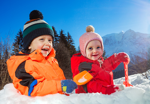Boy with sister girl play standing in snow fortress together over Alps mountains on background