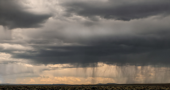 Rain on the horizon with beginnings of sunset showing through under massive dark clouds- foreground is desert with brush sage - mountains in distance - Room for copy