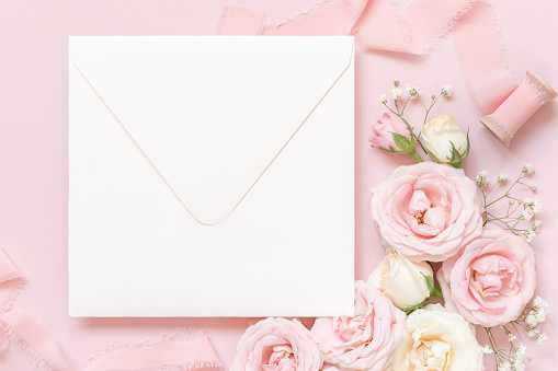 Square envelope between light pink roses and silk ribbons on pink top view,  wedding mockup. Romantic scene with blank envelope and pastel flowers flat lay. Valentines, Spring or Mothers day