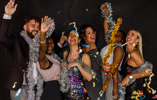 Candid shot of joyful group of diverse, young people, friends, standing on the dance floor, laughing, bonding, having fun playing with shiny tinsel garlands and dancing during a New Year's countdown while colorful confetti are falling down on them.