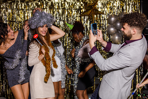 Candid shot of cheerful young women making a hat out of a sparkly tinsel garland, dancing, laughing and having while while their male friend is taking photos with smart phone. More people dancing in the background and having fun celebrating New Year's eve.