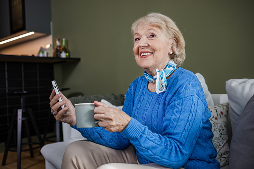 Happy senior woman relaxing in the living room. She is enjoying in her coffee and using smart phone.