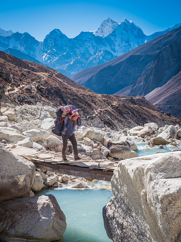 Sherpa porter transporting a heavy load using traditional namlo head band over a wooden footbridge at the edge of the Khumbu glacier, deep in the Himalaya mountains of the Everest National Park, a UNESCO World Heritage Site, Nepal.