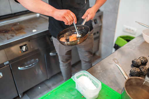 Indoor shot of a busy kitchen where a male cook is working. Male hands are preparing a salmon dish using a frying pan and a long spoon.