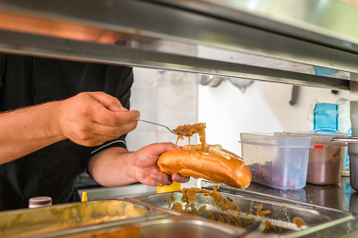 Indoor shot of a male chef preparing a hot dog. He is wearing a black uniform and using a long spoon.