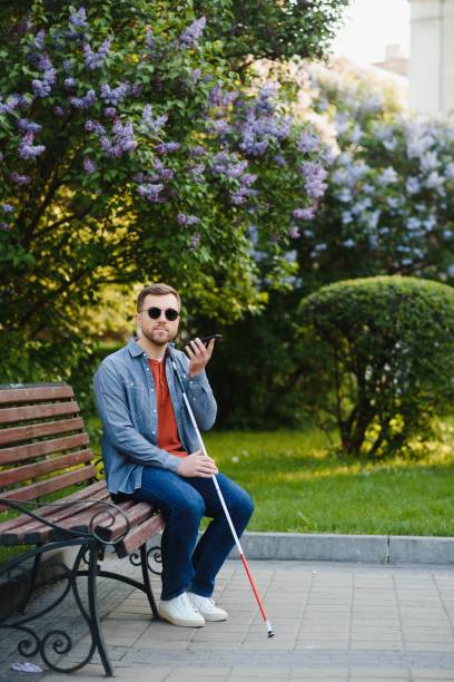istock Visually impaired man with walking stick, sitting on bench in city park. Copy space 1455338663