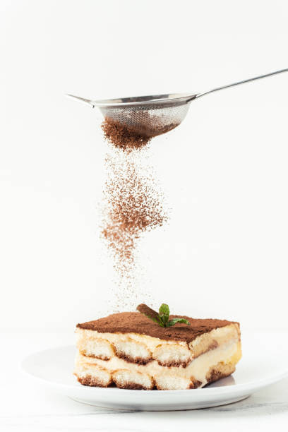 Pastry Chef sprinkles cocoa powder through a sieve on a tiramisu cake decorated with green mint. Making traditional Italian no-baking dessert. White background