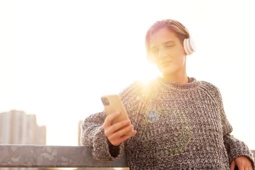 Stylish blonde short haired woman in headphones listening to music outdoors during sunset using playlist in her mobile phone.