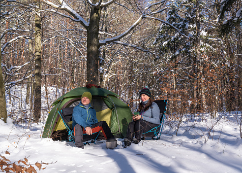 A man and a woman are sitting on chairs in a snowy forest near a tent. Tea preparation. Active life. Joy and satisfaction in life. Frosty air, burning food. Sunny day. A lot of snow.