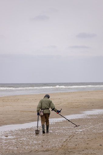 Cotuit, Massachusetts, USA - February 6, 2021-  A bearded man wearing boots and rubber waders uses a metal detector in the shallow water of Loop Beach on a cold February afternoon.