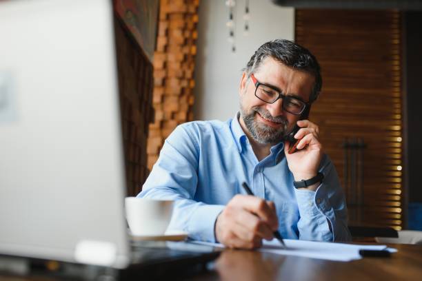 istock Positive senior bearded man with grey hair drinking coffee and using laptop at cafe, copy space. Stylish aged businessman in burgundy jacket enjoying his tea while working online. 1455347029