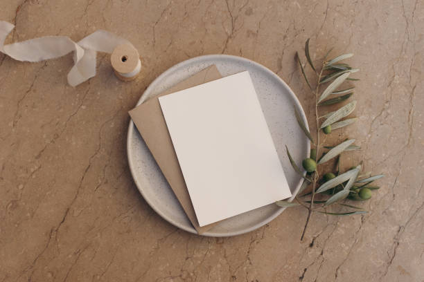 istock Blank greeting card, invitation and envelope mockup on ceramic plate. Beige marble stone tiles background with olive tree branch, ribbon. Summer wedding composition, office supply. Flatlay, top view. 1455349302