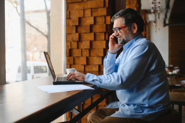 istock Positive senior bearded man with grey hair drinking coffee and using laptop at cafe, copy space. Stylish aged businessman in burgundy jacket enjoying his tea while working online. 1455349363